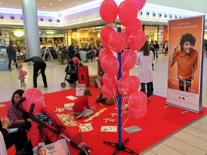 Bambini che giocano su un tappeto rosso in un centro commerciale, circondati da palloncini rosa e materiali promozionali per la prevenzione oncologica.