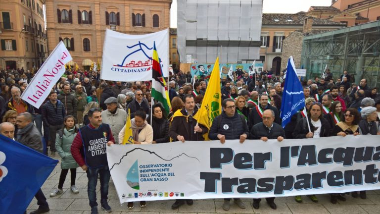 Manifestazione per la difesa dell’acqua pubblica con striscioni, bandiere e folla in piazza; in evidenza lo slogan “Per l’Acqua Trasparente”.