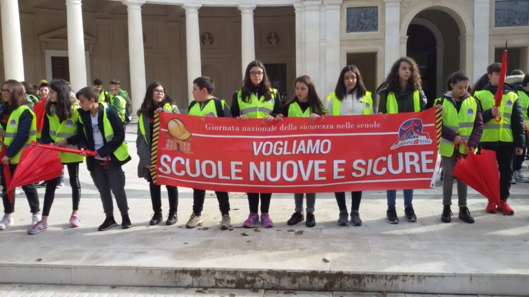 Studenti con giubbotti riflettenti tengono uno striscione rosso con scritto "Vogliamo scuole nuove e sicure" durante una manifestazione scolastica.