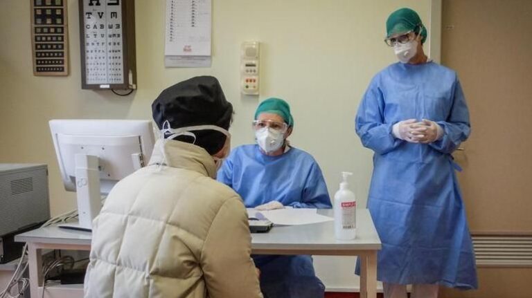 Visita medica in ambulatorio con due dottoresse in camice e mascherina che parlano con una paziente seduta alla scrivania.