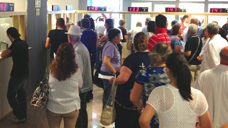 Persone in fila agli sportelli di un ufficio pubblico con display numerici luminosi che segnalano i turni di attesa.