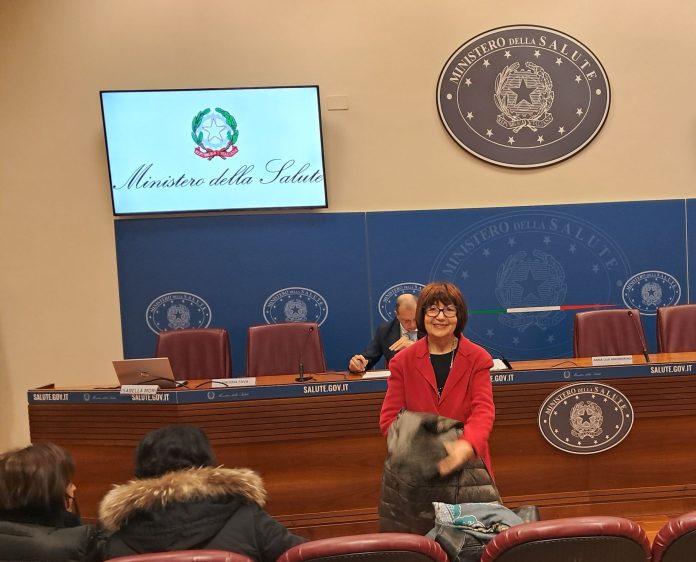 Donna sorridente in primo piano in una sala conferenze del Ministero della Salute, con schermo e logo istituzionale ben visibili.